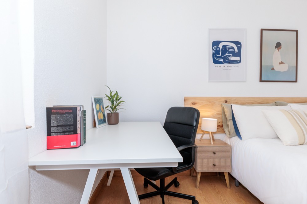 Escritorio con libros, silla de oficina y lámpara, junto a la cama